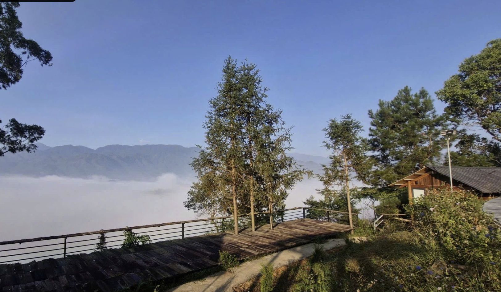 Una terraza de madera con barandilla metálica que da a un paisaje montañoso con zona boscosa y un pequeño edificio.