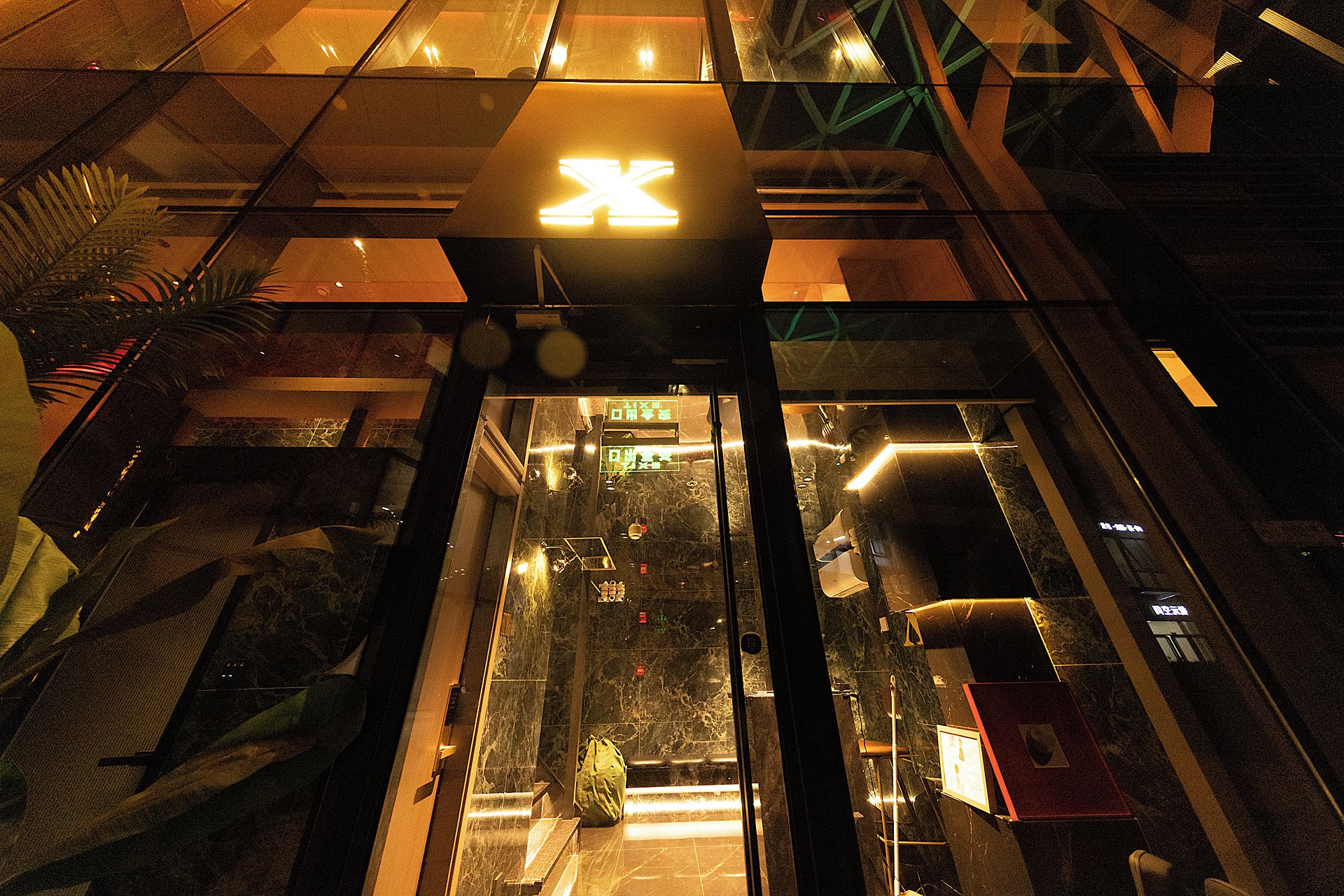 An interior view of a modern building with a glass elevator shaft, reflective surfaces, and a geometric ceiling design.
