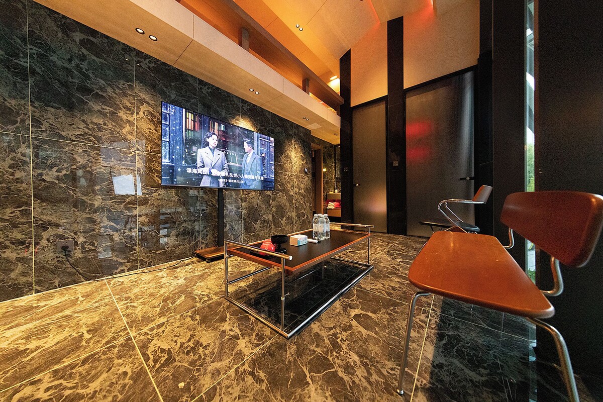 A modern lobby with a large flat-screen TV mounted on a marble wall, a reception desk, and two red chairs.