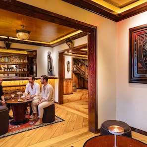 A bar area with wooden flooring, a round table with two people seated, a bar with various bottles, and a staircase leading to another level. - Hotel Parque Mexico - Petit (Mexico City)