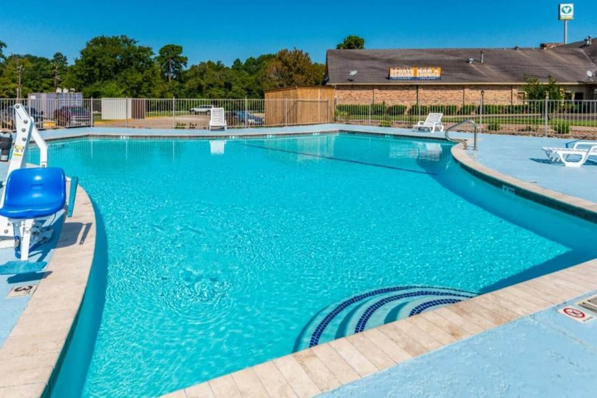 A clear blue swimming pool with a wooden deck, lounge chairs, and a fenced area with a building in the background.