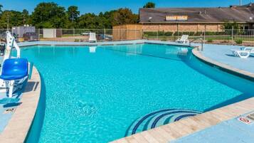 A clear blue swimming pool with a wooden deck, lounge chairs, and a fenced area with a building in the background.