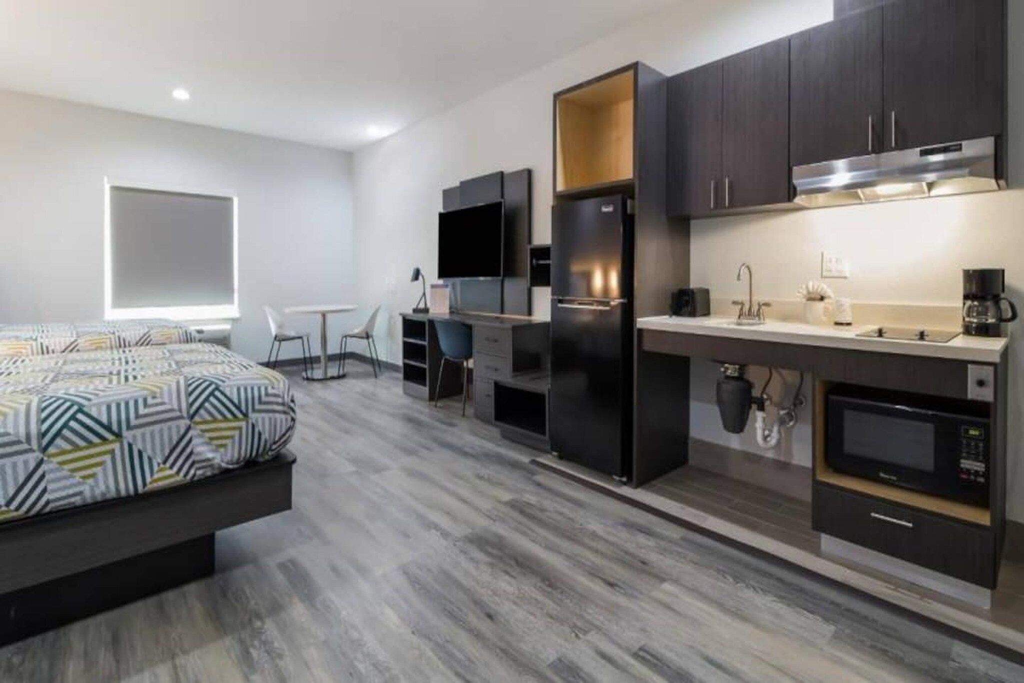 A modern hotel room with a bed, a desk, a flat-screen TV, a refrigerator, a microwave, a coffee maker, and a window.
