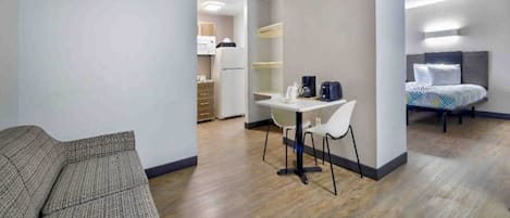 A modern hotel room with a sofa, a dining table with chairs, a kitchenette, and a bed with a headboard.