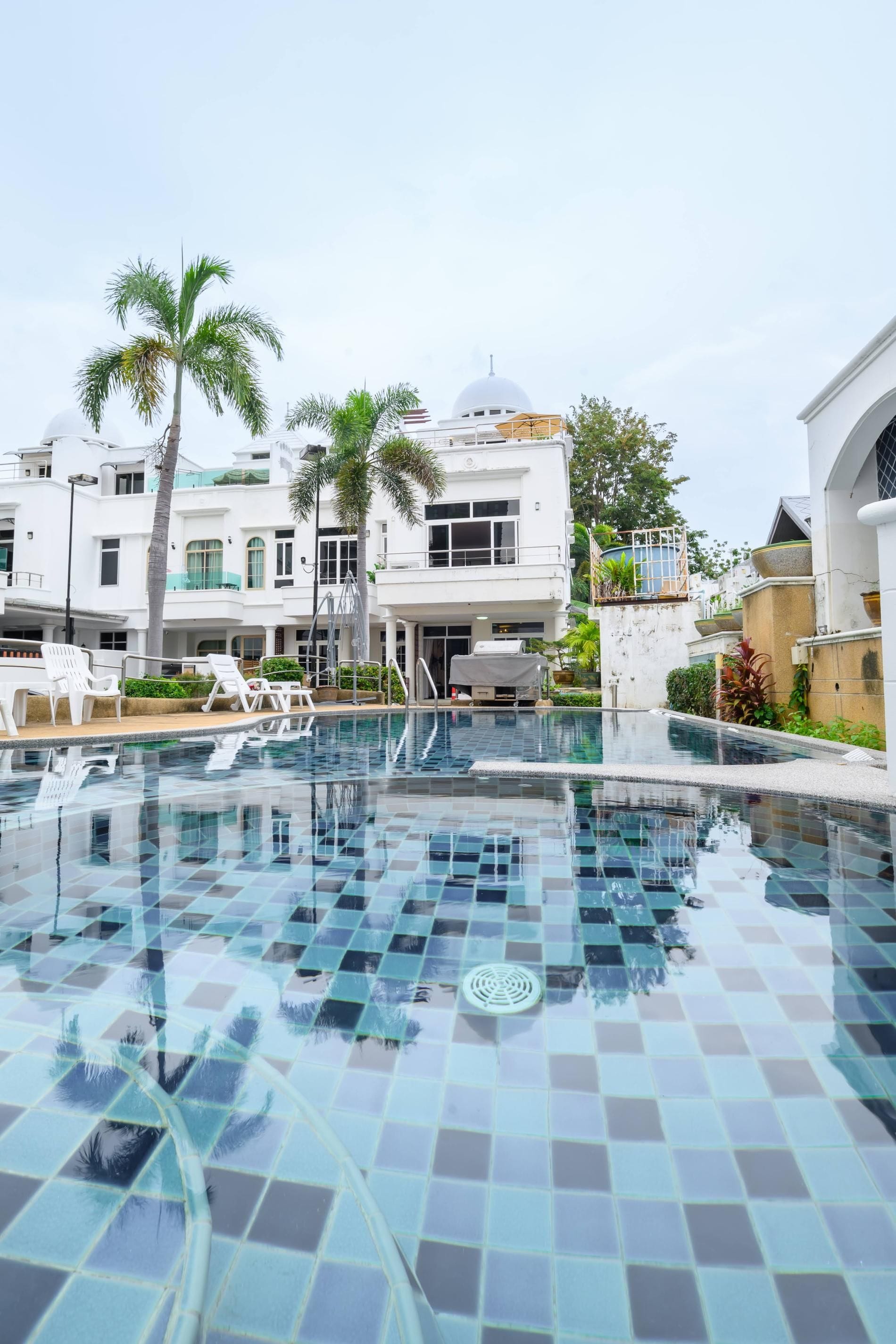 Ein Pool mit Kachelboden, umgeben von Palmen und einem Gebäude mit weißer Fassade.
