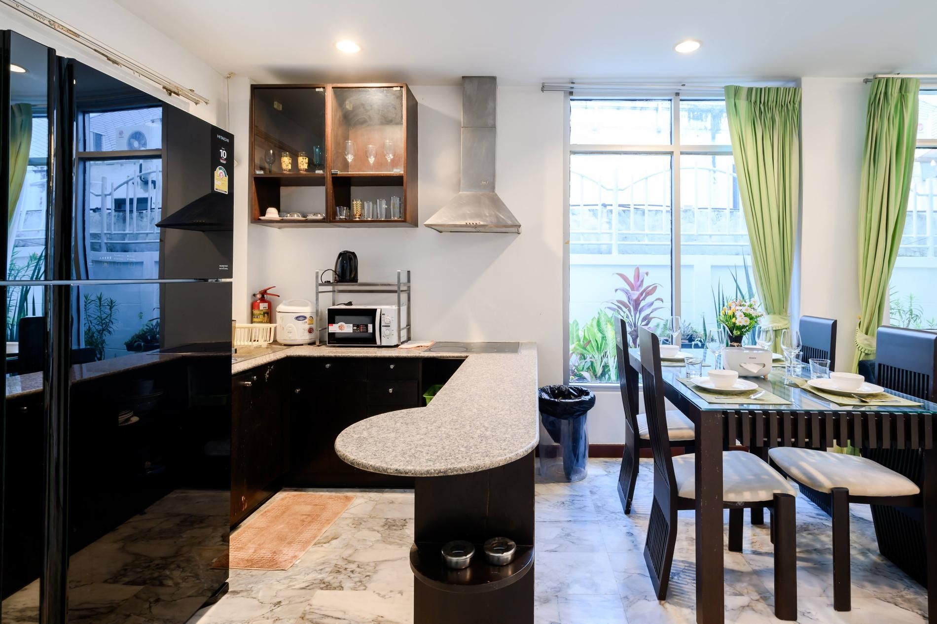 Een moderne keuken met een ingebouwde magnetron, een koffiezetapparaat en een gasfornuis. Er is een eethoek met een tafel voor twee personen, stoelen en een raam met gordijnen.