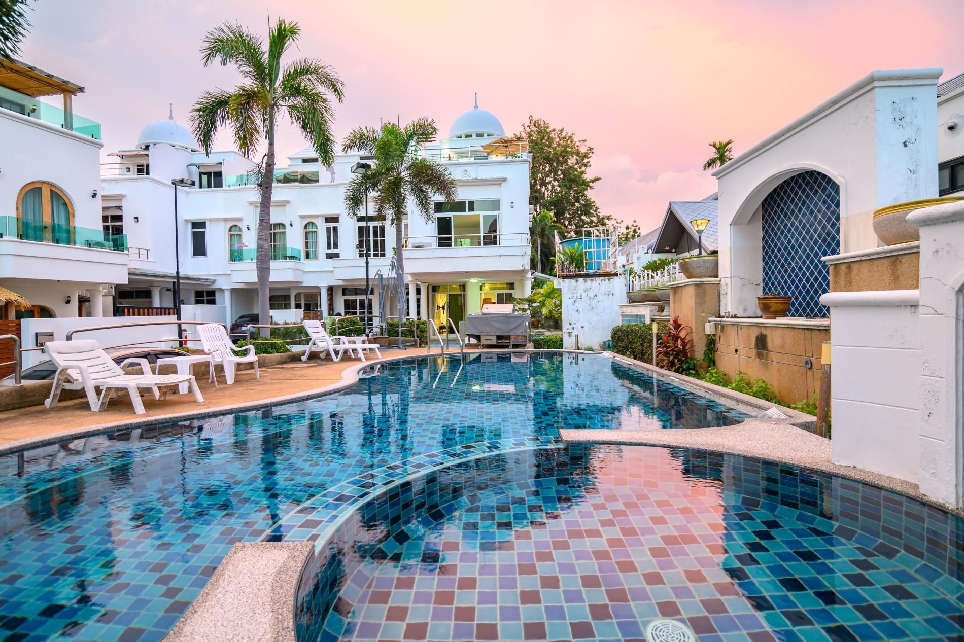 A swimming pool with tiled flooring, surrounded by lounge chairs and palm trees.