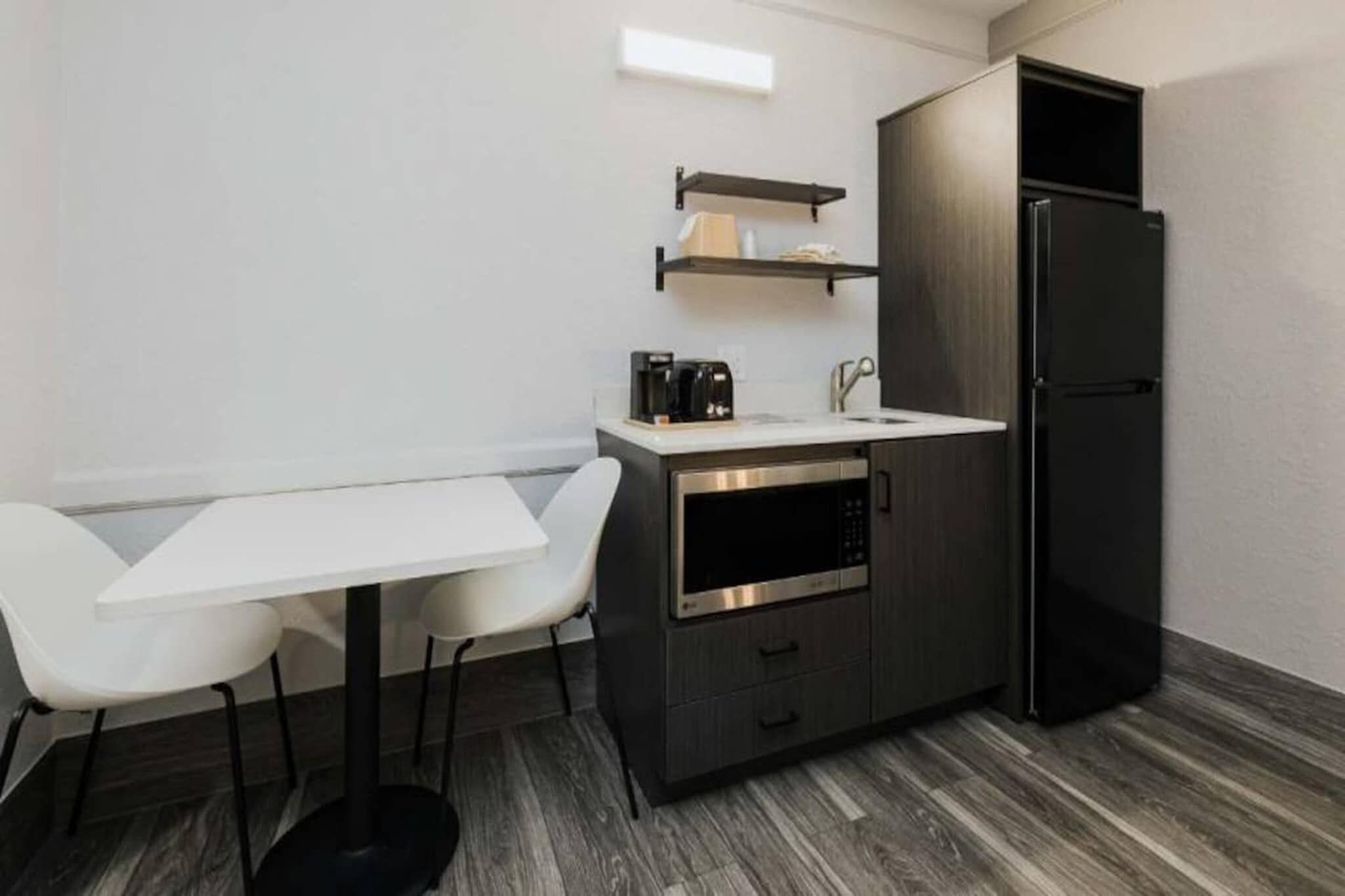 A compact kitchenette with a white countertop, a black refrigerator, and a built-in oven. There are two white chairs and a small shelf above the counter.