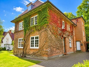 A two-story brick house with a sloped roof, covered partially by green and red foliage, and a paved driveway. - Holiday Apartment With 1 Bedroom (Wyk auf Foehr)