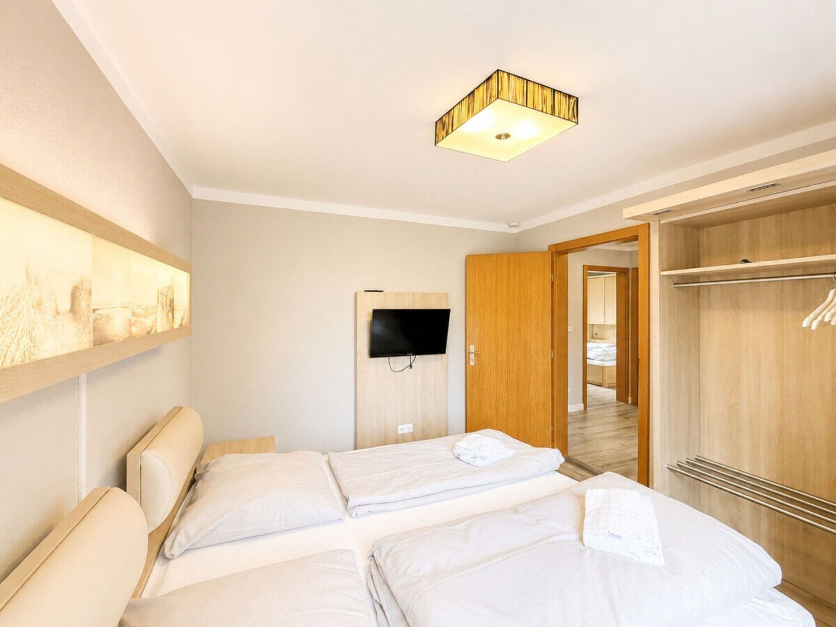 Een hotelkamer met twee bedden, een wandtelevisie, een houten kledingkast en een gang naar een andere kamer.