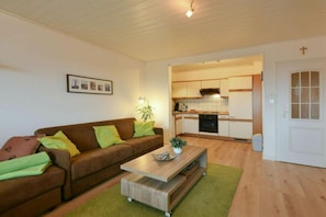 A living room with a brown sofa, a coffee table, and a green rug. There is a kitchen visible in the background with white cabinets and a stove. - Apartment 17 in Wangerooge (Wangerooge)