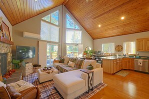 A spacious living area with a high ceiling, large windows, a fireplace, and a kitchen with wooden cabinets. - Cozy Chalet on Golf Course w/ Lake Cushman Access (Hoodsport)