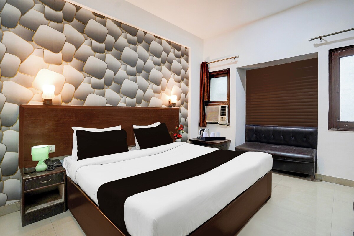 A hotel room with a large bed, a headboard with a geometric pattern, a bedside table with a lamp, a small table with a kettle and cups, a seating area with a sofa, and a window with blinds.