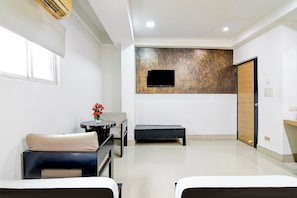Habitación de hotel con dos camas, una pequeña mesa con un jarrón de flores, un banco, un televisor de pantalla plana montado en una pared con textura y una puerta de madera.