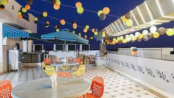 Un espacio de comedor al aire libre con mesas y sillas, una zona de estar cubierta y un techo decorativo con elementos colgantes.