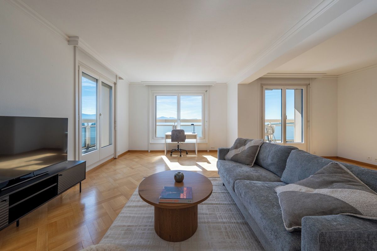 Amplia sala de estar con un sofá grande, una mesa de centro y un televisor. Hay ventanas grandes que ofrecen vistas al exterior.