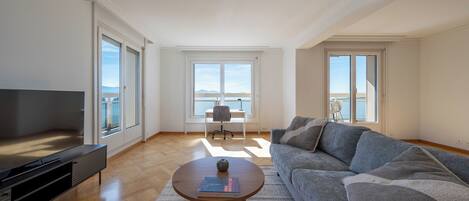 A spacious living room with a large sofa, a coffee table, and a television. There are large windows offering a view of the outdoors.