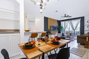 Cozinha moderna com armários brancos, sala de jantar com mesa de madeira e sala de estar com sofá e cortinas.
