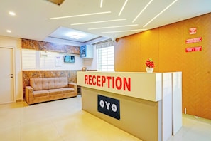 Un área de recepción con un mostrador identificado con 'RECEPTION' y 'OYO'. Se observa un sofá al fondo, un calendario colgado en la pared y un cartel de 'NO FUMAR'.