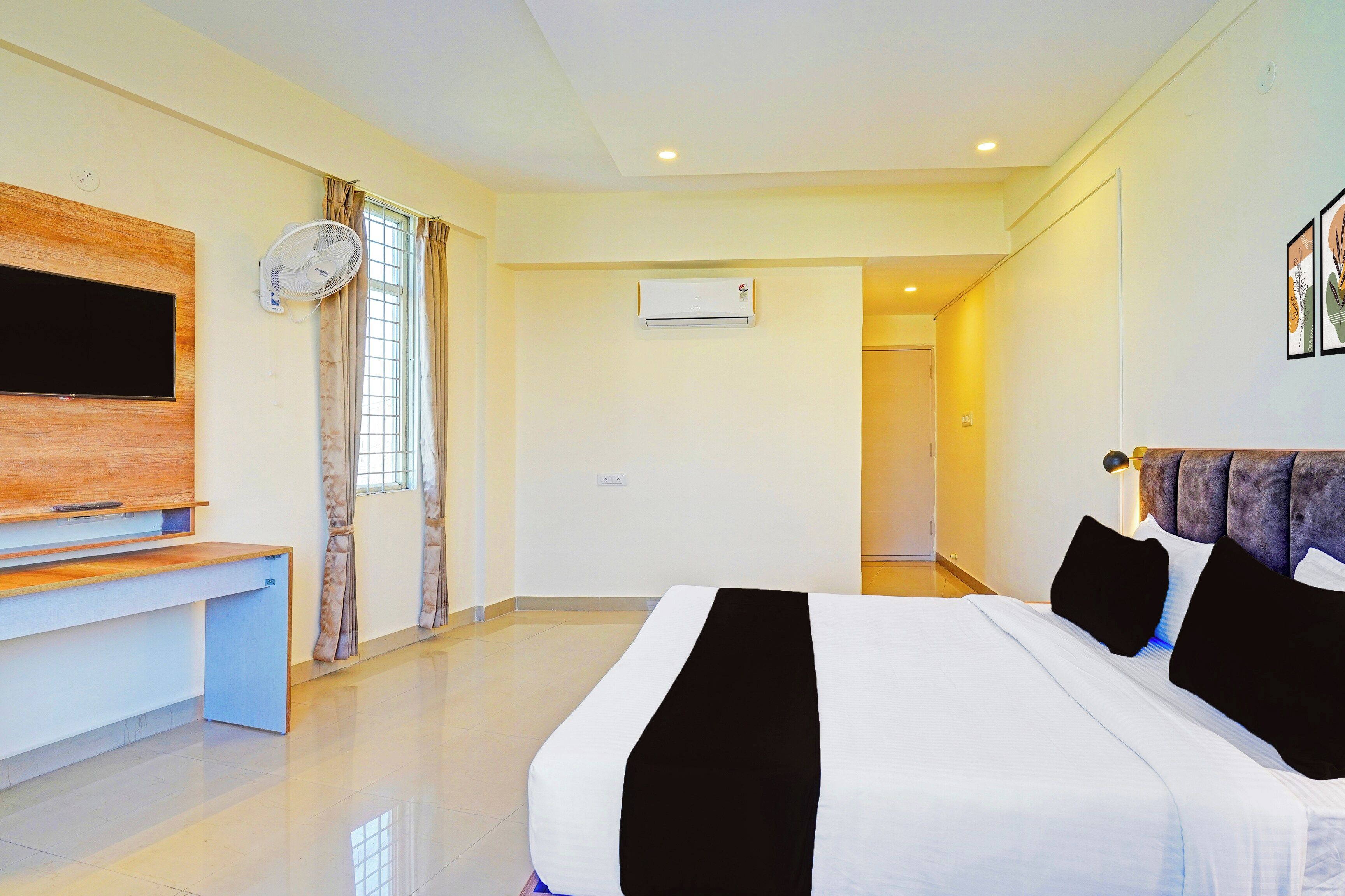 A hotel room with a bed, a desk, a flat-screen TV, an air conditioning unit, and a door leading to another room.