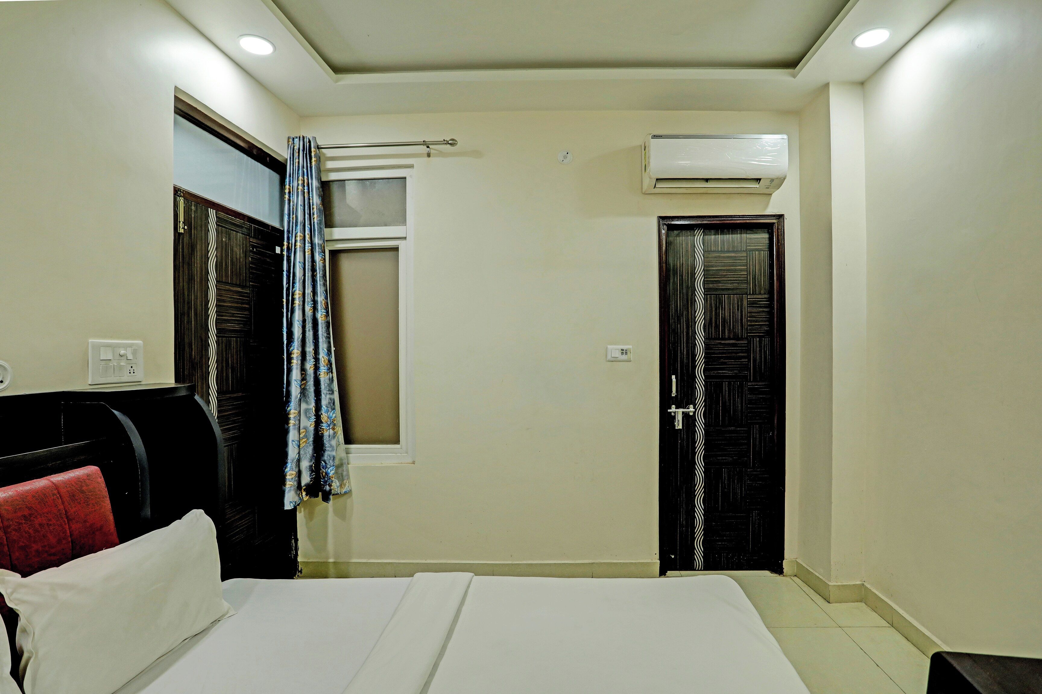 Habitación de hotel con cama, ventana con cortinas, equipo de aire acondicionado y puerta con diseño decorativo.