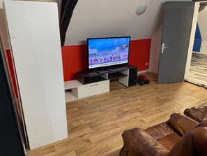 Ein Zimmer mit Fernseher auf einem Standfuß, Holzboden, braunem Ledersofa und einer grauen Tür.