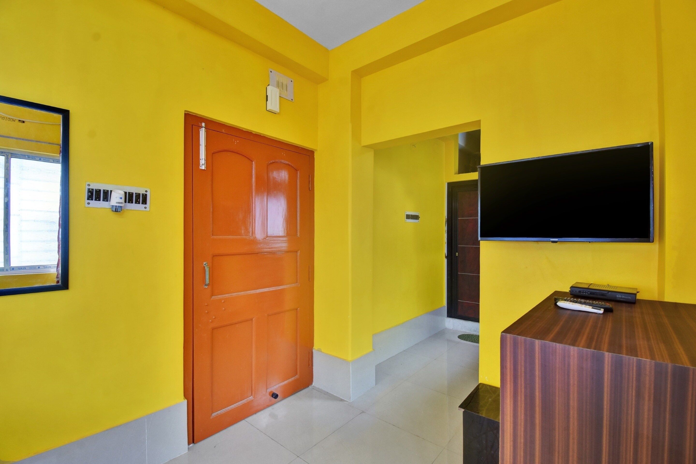 A room with yellow walls, a wooden door, a flat-screen TV mounted on the wall, and a small table with a remote control.