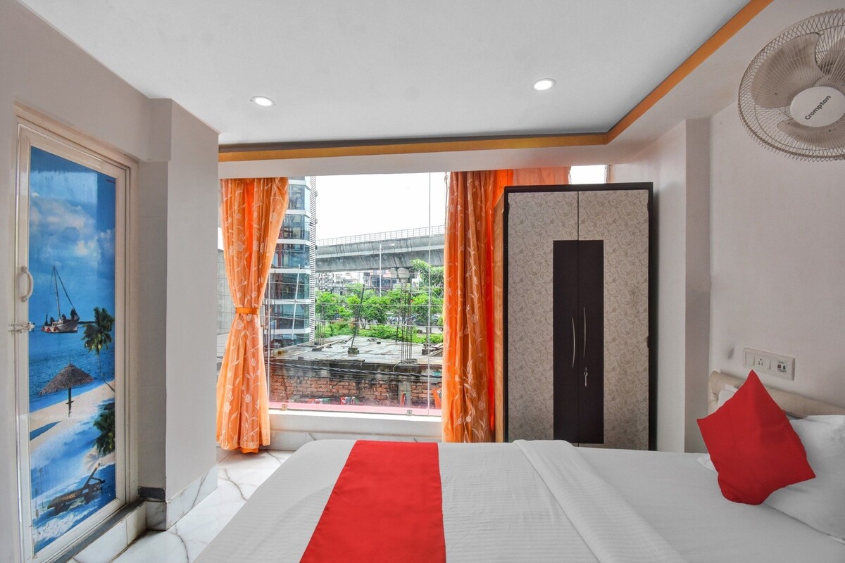 A hotel room with a bed, a window with curtains, a view of a cityscape, and a door with a painting.