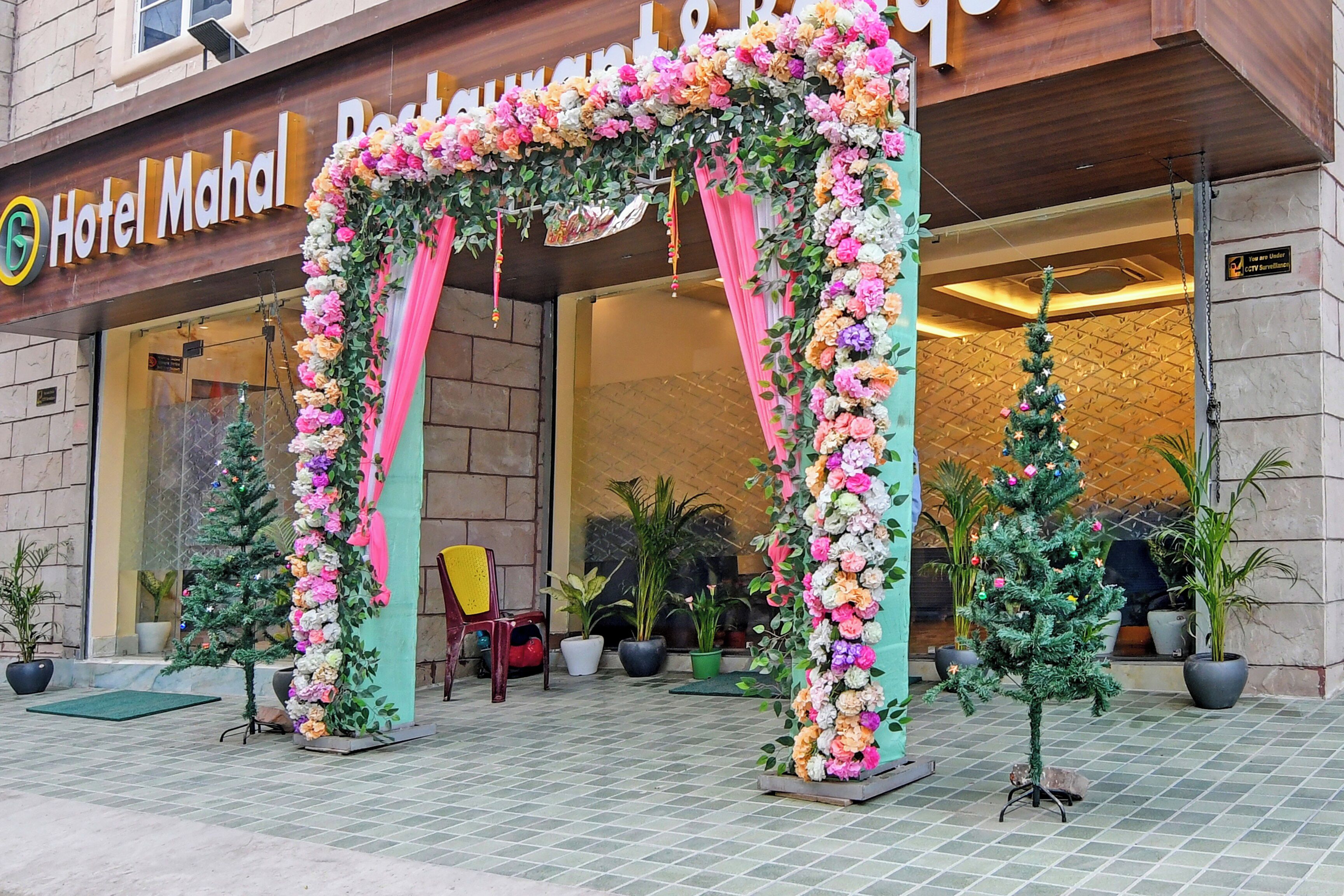 A hotel entrance with a floral archway, potted plants, and a CCTV surveillance sign.