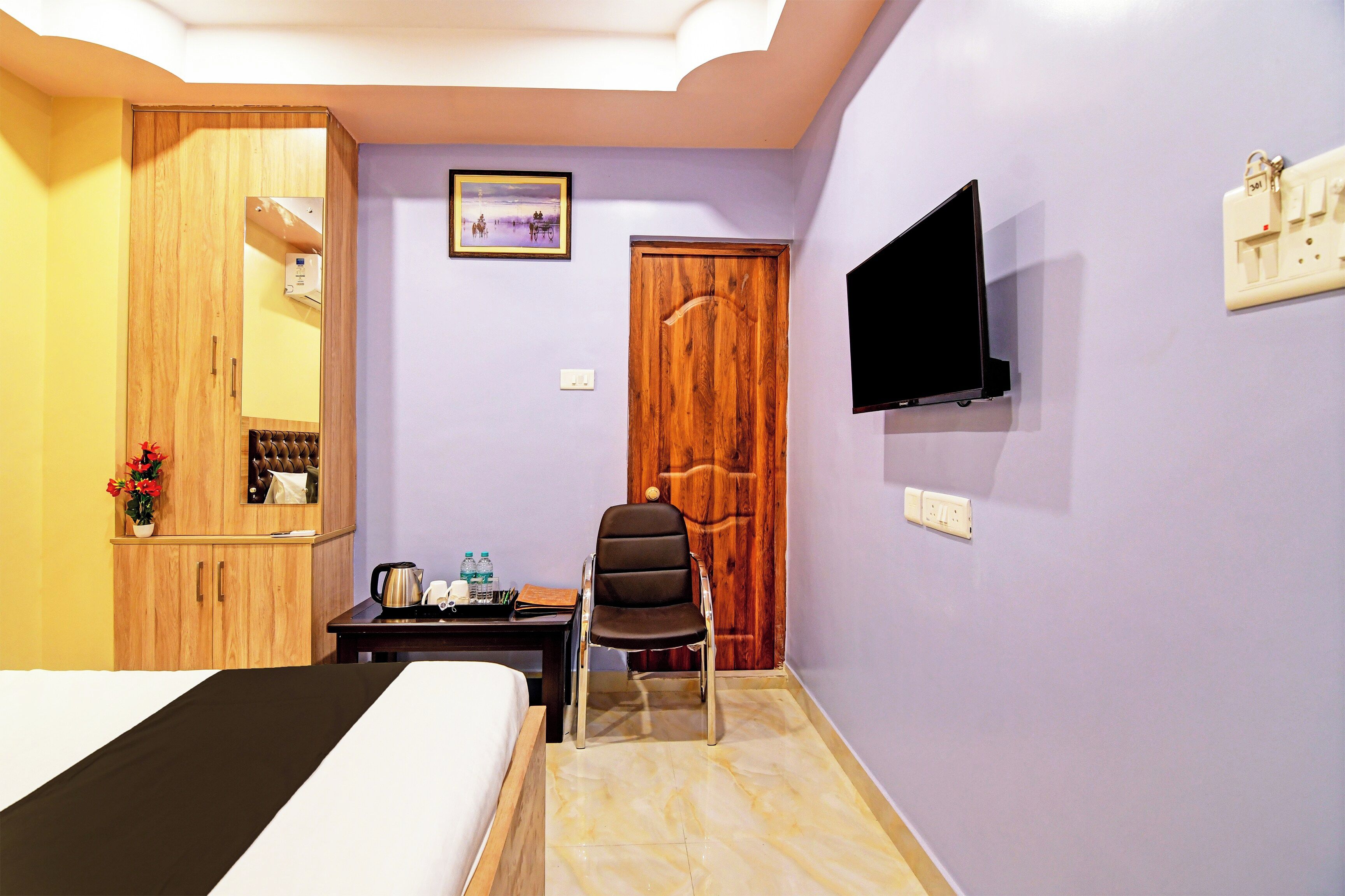 A hotel room with a bed, a wooden wardrobe, a desk with a chair, a television mounted on the wall, and a door.