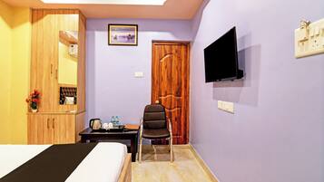 A hotel room with a bed, a wooden wardrobe, a desk with a chair, a television mounted on the wall, and a door.