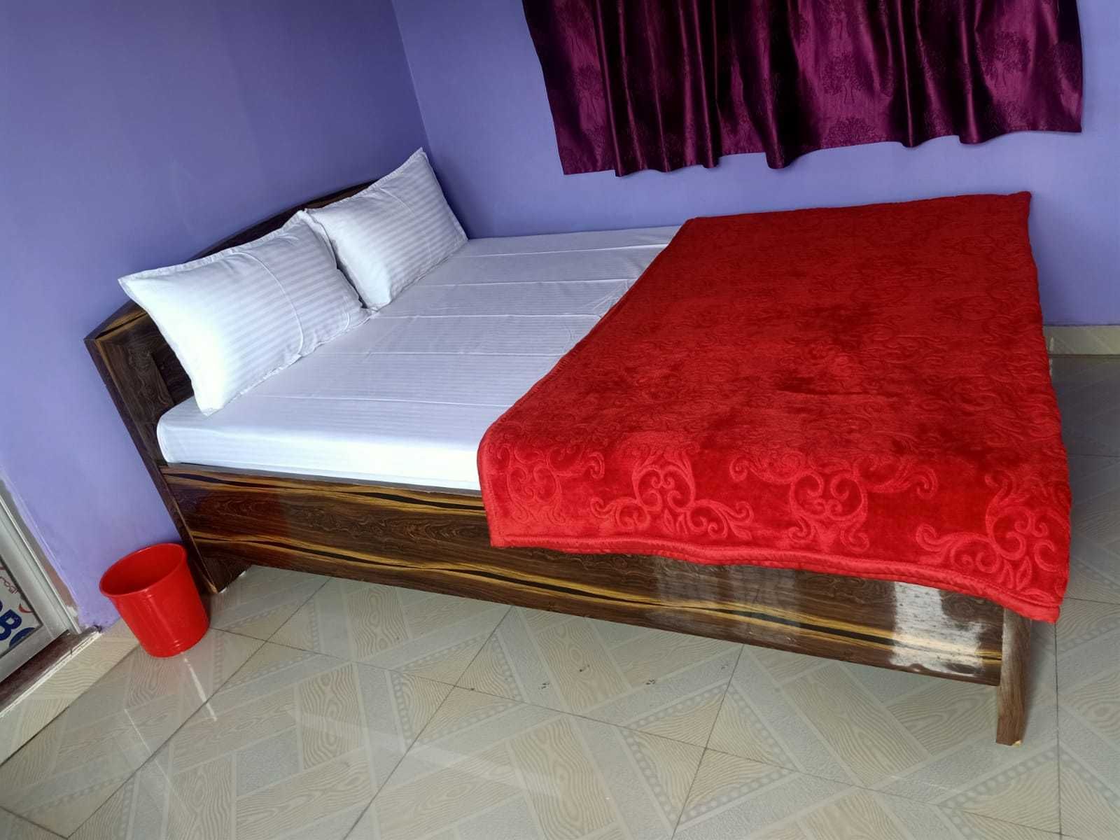 Un lit simple avec un matelas blanc et une couverture rouge, une tête de lit en bois, un seau rouge et un rideau violet.