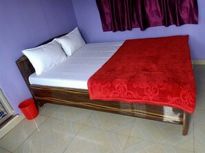 A single bed with a white mattress and a red blanket, a wooden headboard, a red bucket, and a purple curtain.
