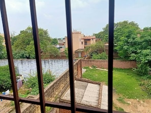 A view through a window with a metal frame, featuring a brick wall, a grassy area, and buildings in the background.