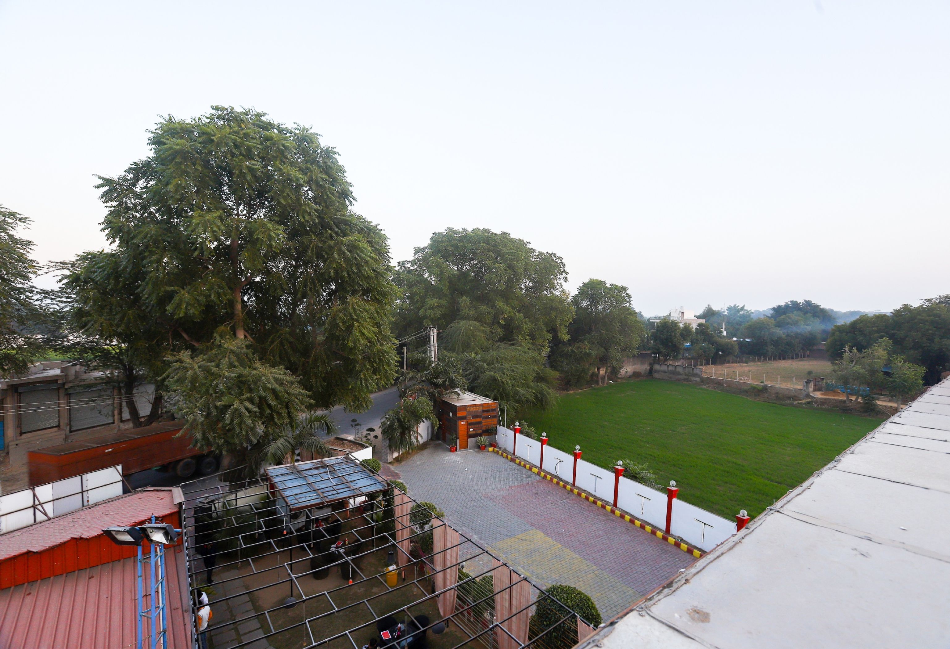 A view of a building with a paved area, a fenced sports field, and trees in the background.