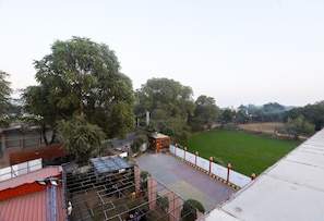 Vista de un edificio con una zona pavimentada, un campo deportivo cercado y árboles al fondo.
