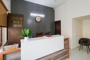 Una mesa de recepción con un reloj, un cartel que indica horarios de registro de entrada y salida, y un aviso de GST 12% adicional.