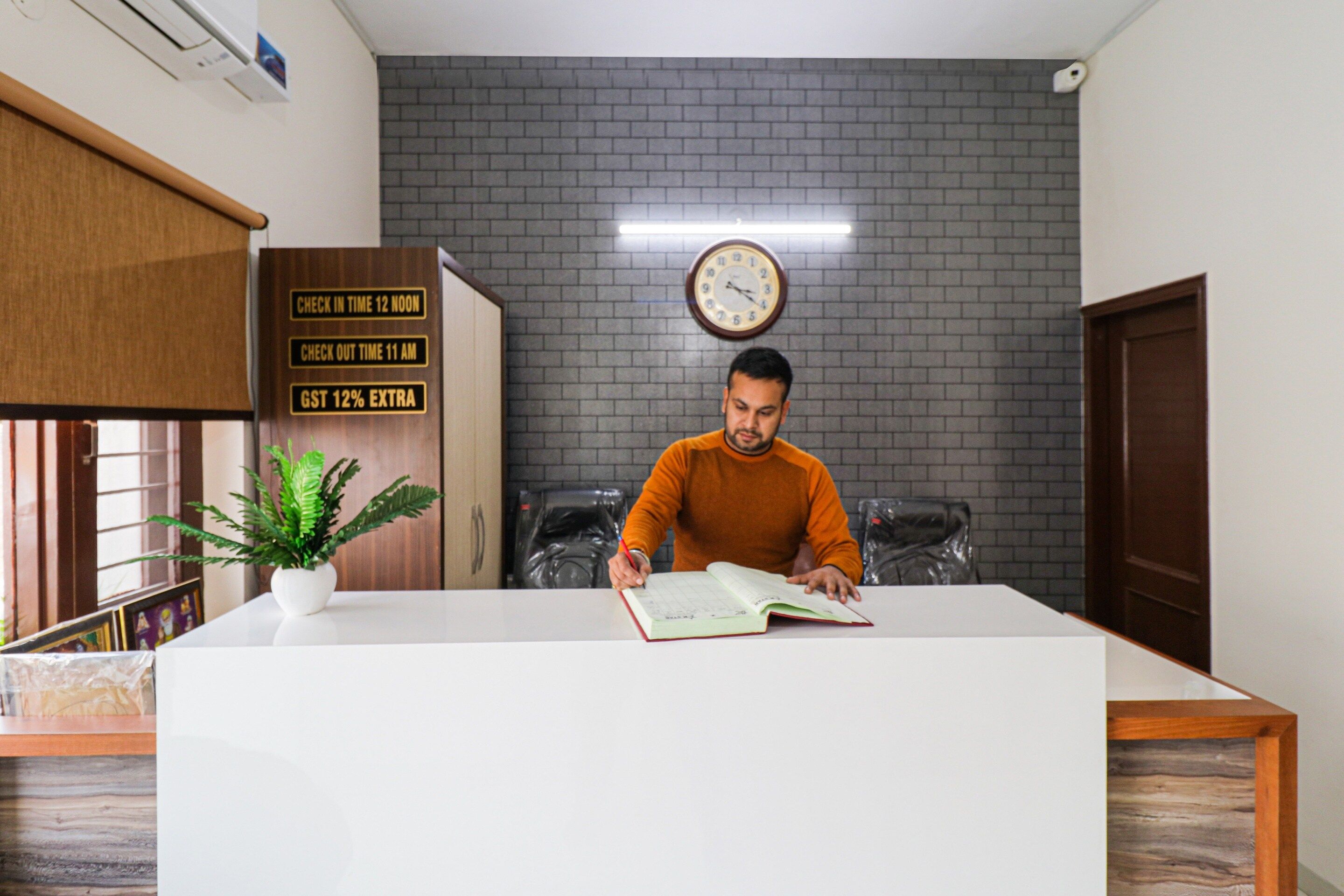 Un hombre sentado en un escritorio con un libro, un reloj en la pared y un cartel que indica horarios de registro de entrada y salida.