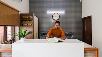 Un hombre sentado en un escritorio con un libro, un reloj en la pared y un cartel que indica horarios de registro de entrada y salida.