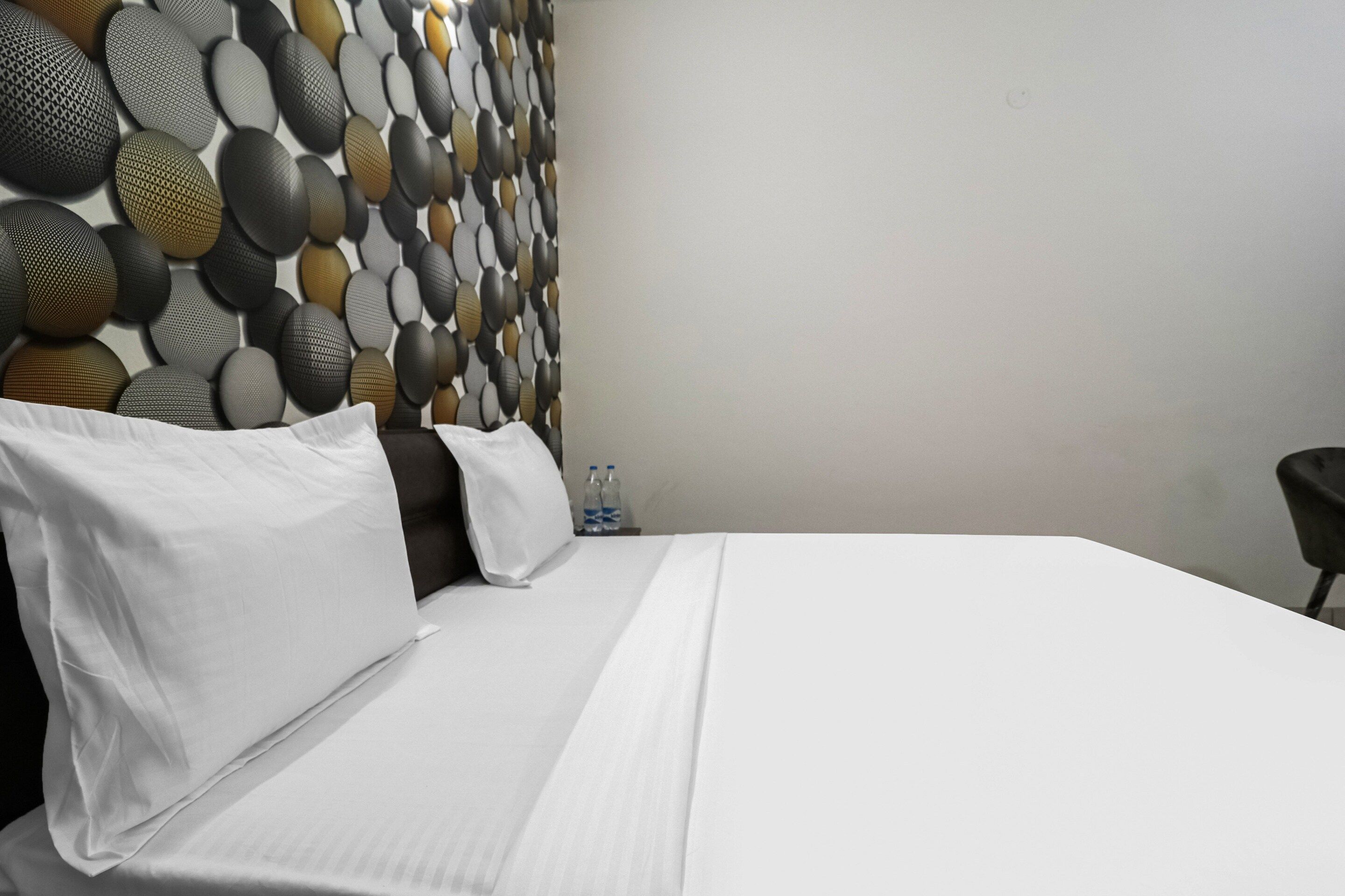 A hotel room with a neatly made bed, a headboard with a textured pattern, and a plain wall.
