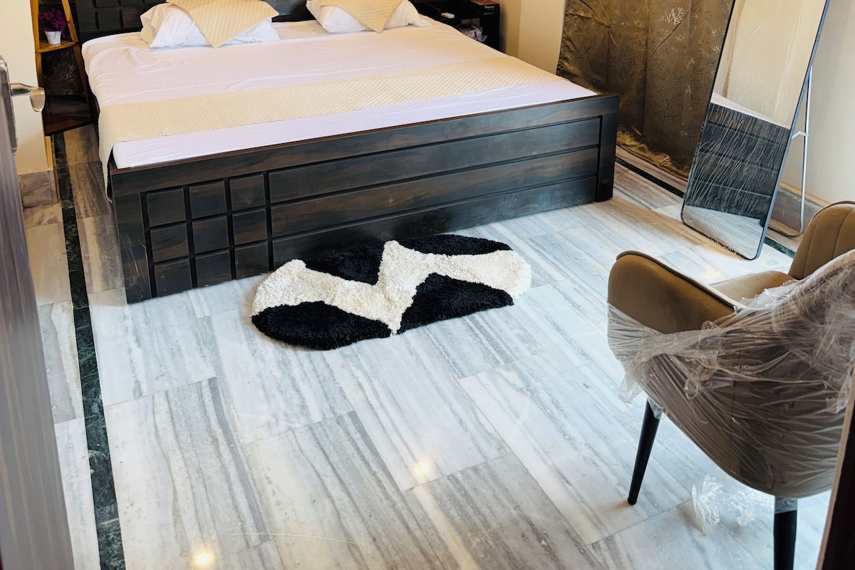 A hotel room with a bed, a nightstand, a lamp, a chair, a mirror, and a bathroom with a shower curtain.