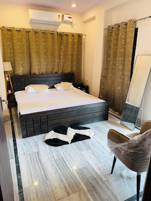 A hotel room with a bed, a nightstand, a lamp, a chair, a mirror, and a bathroom with a shower curtain. - Angan E Kashi (Varanasi)