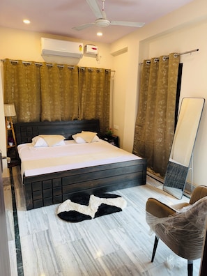 A hotel room with a bed, a ceiling fan, an air conditioning unit, a large mirror, and a patterned curtain. - Angan E Kashi (Varanasi)