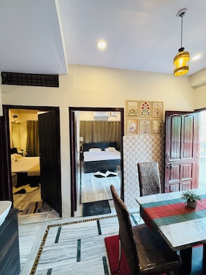 A hotel room with a bed, a bathroom, a dining area with a table and chairs, and a hallway leading to another room. - Angan E Kashi (Varanasi)
