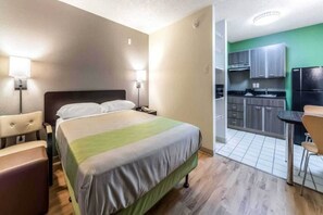 A hotel room with a bed, bedside table, lamp, and a kitchenette area with cabinets and a refrigerator. - 1 Queen Bed | Smoking (Dallas)