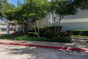 A parking area with a red curb indicating no parking, surrounded by greenery and a building with a white facade. - 1 Queen Bed | Non-Smoking (Dallas)
