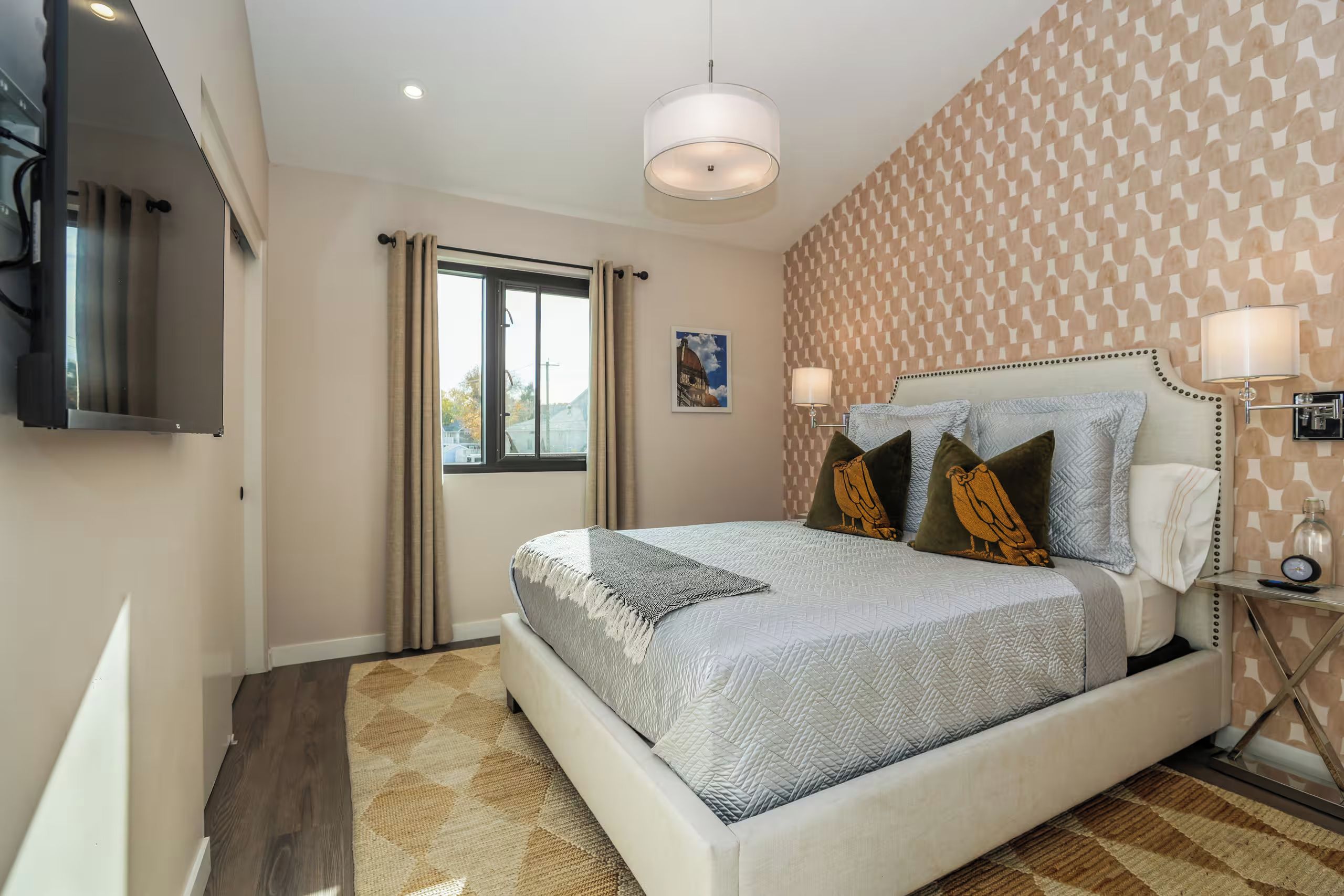 A hotel room with a bed, a TV, a window with curtains, and patterned wallpaper.