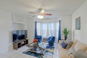 A living room with a ceiling fan, a flat-screen TV, a coffee table with a vase of flowers, a sofa with cushions, and a potted plant. - Lovely Merrimack Valley Home Near Local Eats (Lawrence)