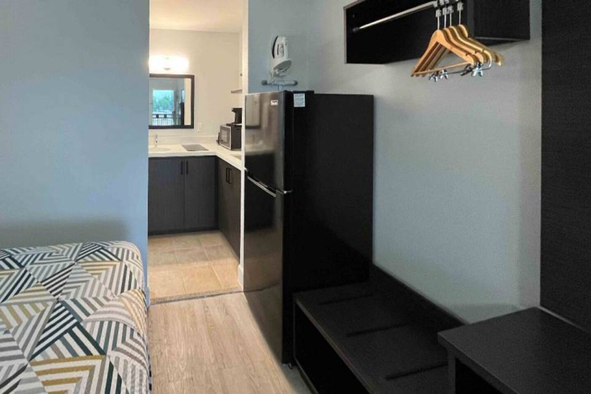 A compact room with a bed, a small kitchenette, a black TV stand, and a wooden hanger rack.