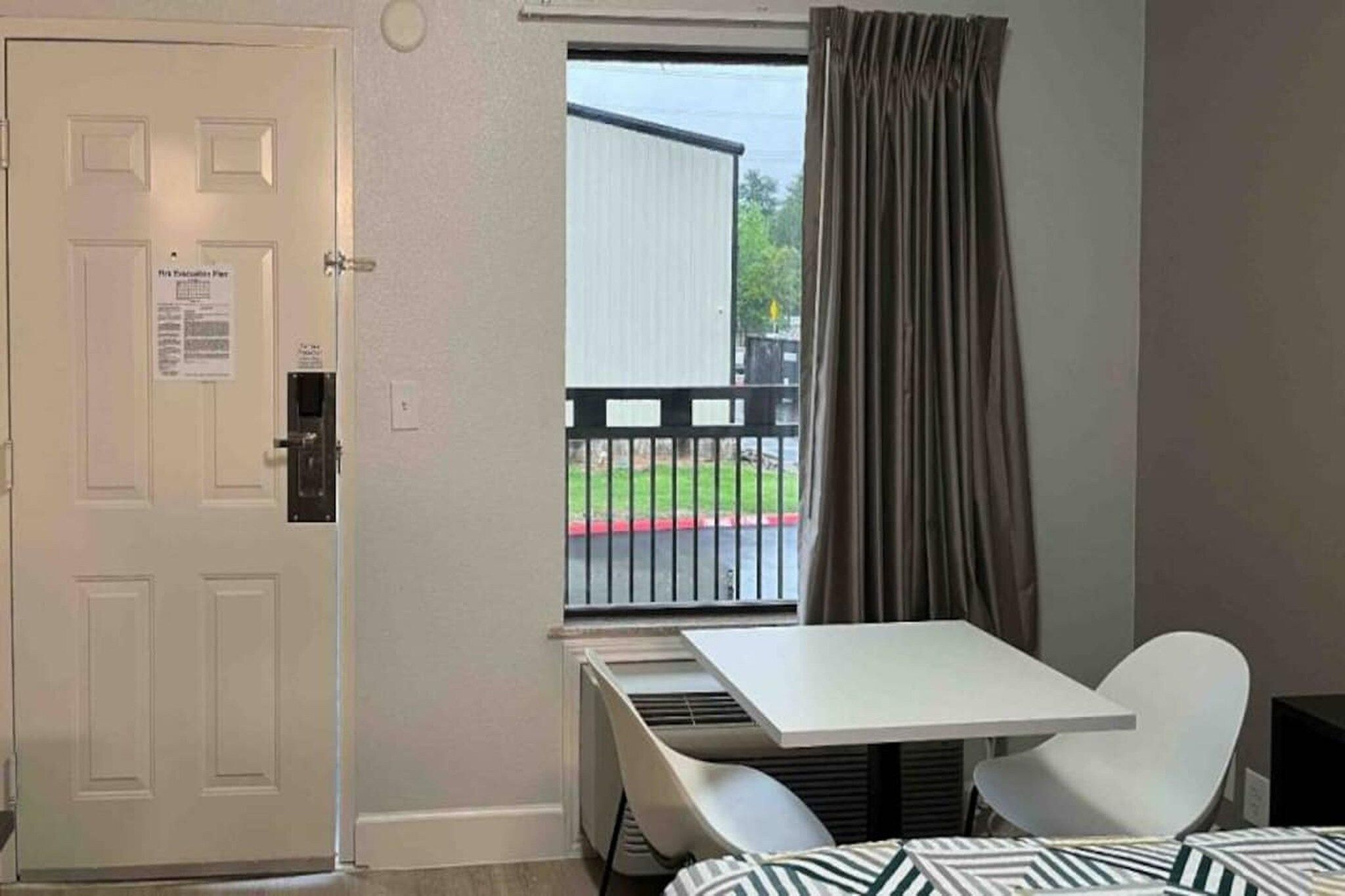 A hotel room with a white door, a window with a view of a balcony, a small table, and a white chair.